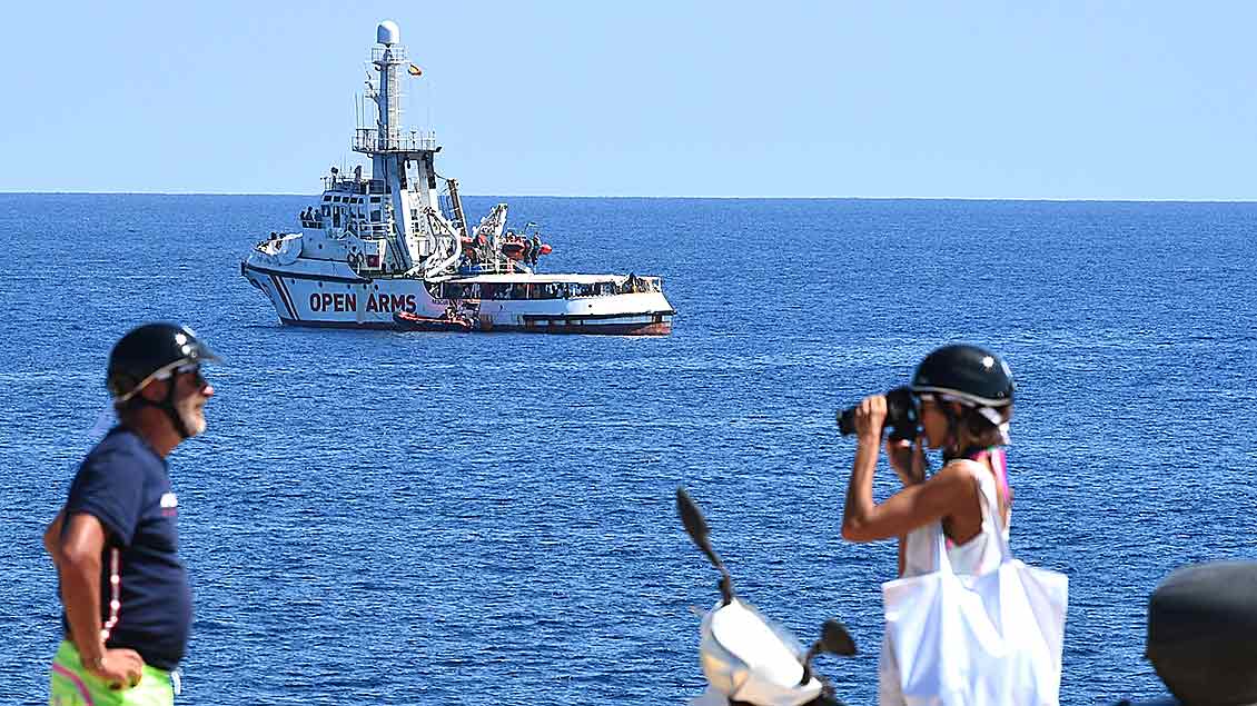 Eine junge Frau fotografiert einen Mann während im Hintergrund auf dem Meer das Rettungssschiff Opern Arms zu sehen ist.
